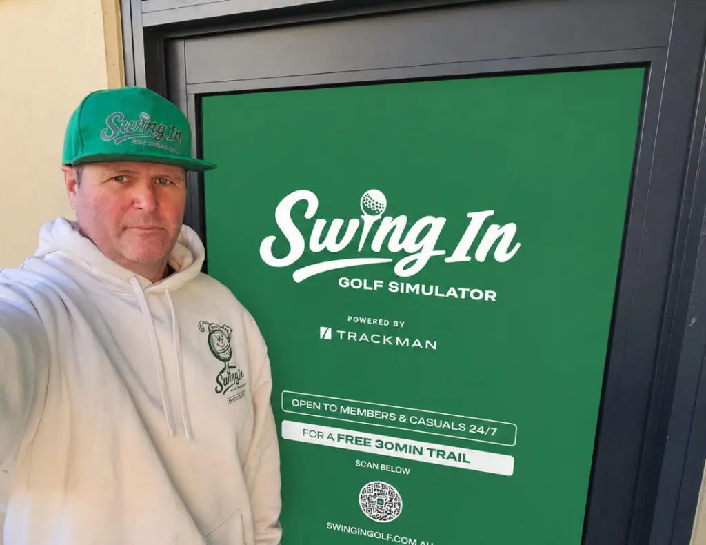 Man in a white hoodie and green cap posing beside a large green Swing In Golf Simulator sign, with TrackMan branding and a note about a free 30-minute trail.