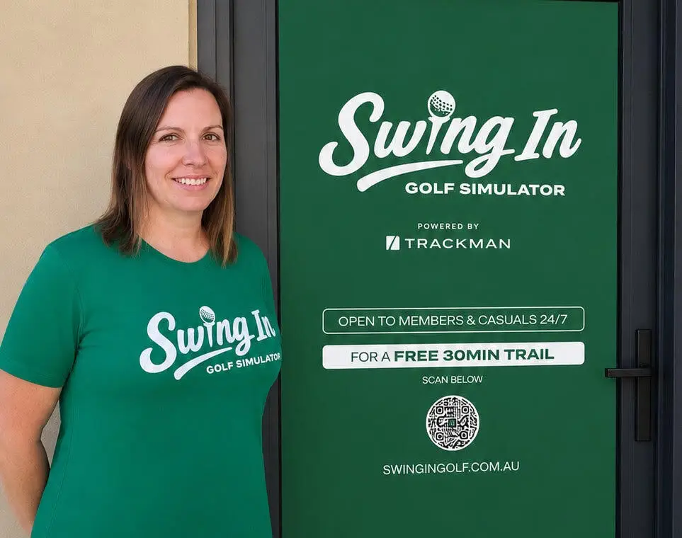 Smiling woman in a green 'Swing In Golf Simulator' shirt stands next to a branded green door with the logo.
